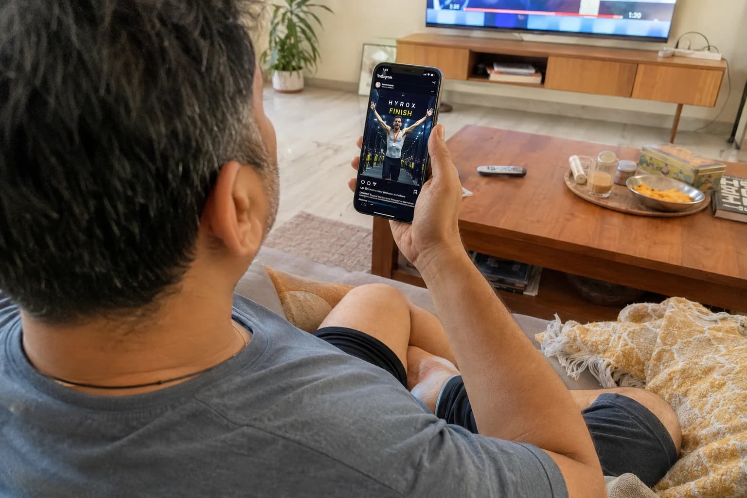 Someone scrolling Instagram past Hyrox finish line photos while sitting on a couch — the gap between aspiration and reality