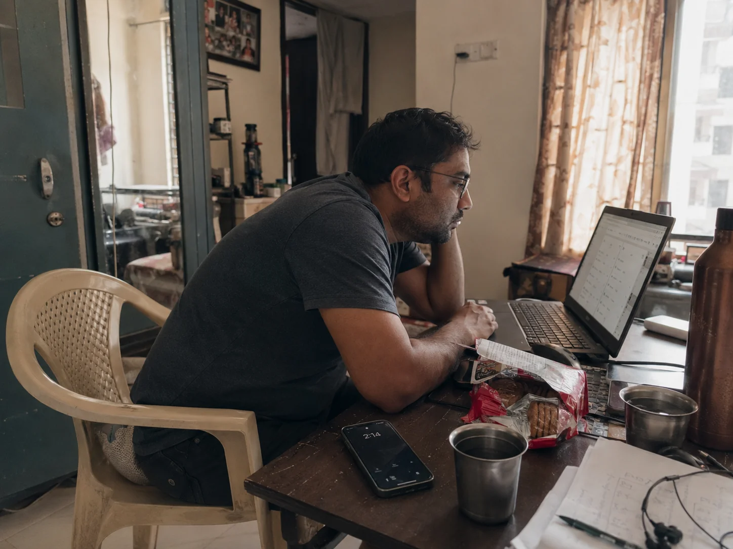 Indian man hunched at WFH desk — laptop, empty chai cups, biscuit packet, phone showing 2pm