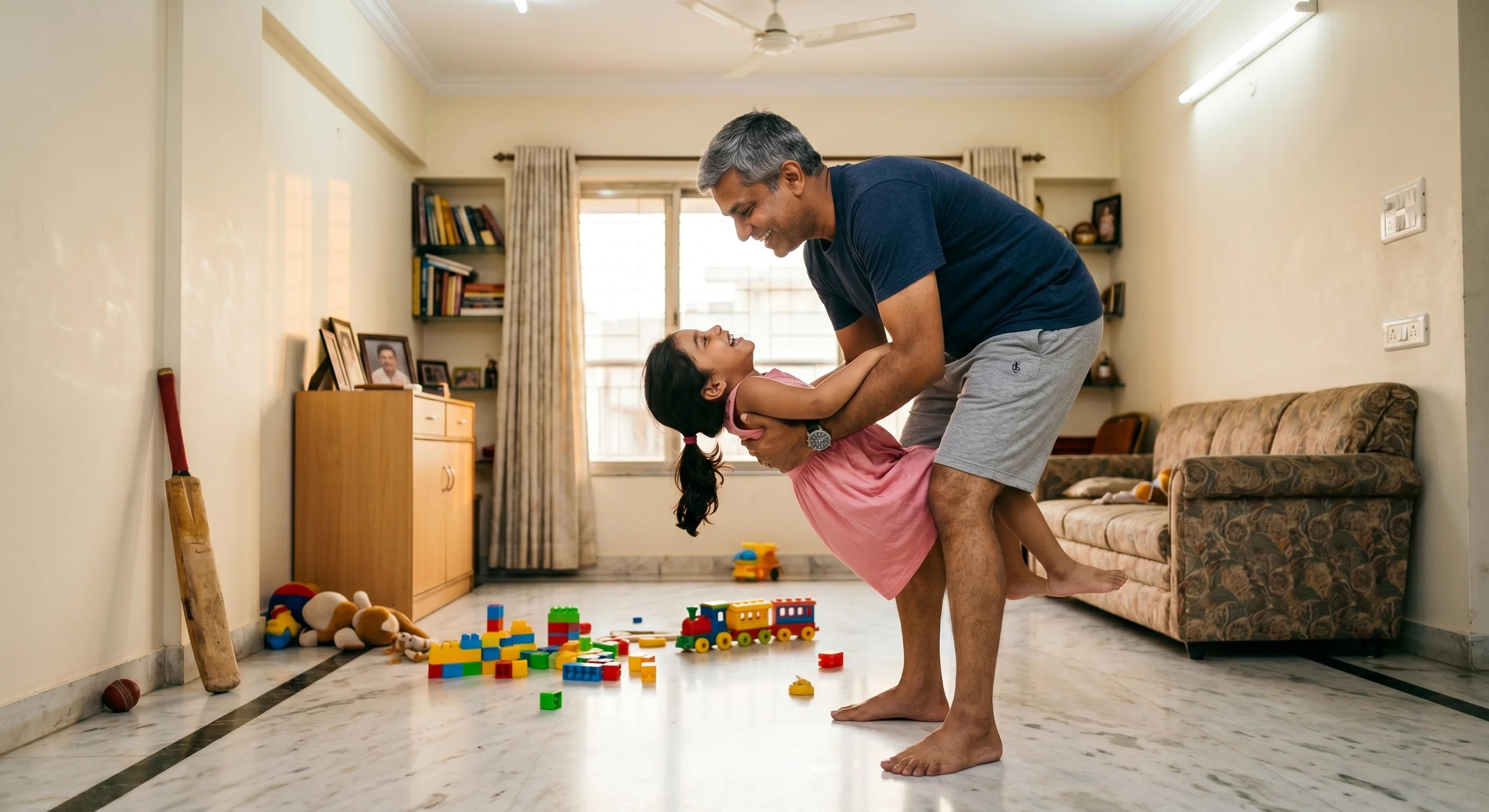Indian father scooping up his daughter from the floor effortlessly — toys scattered, living room, evening light
