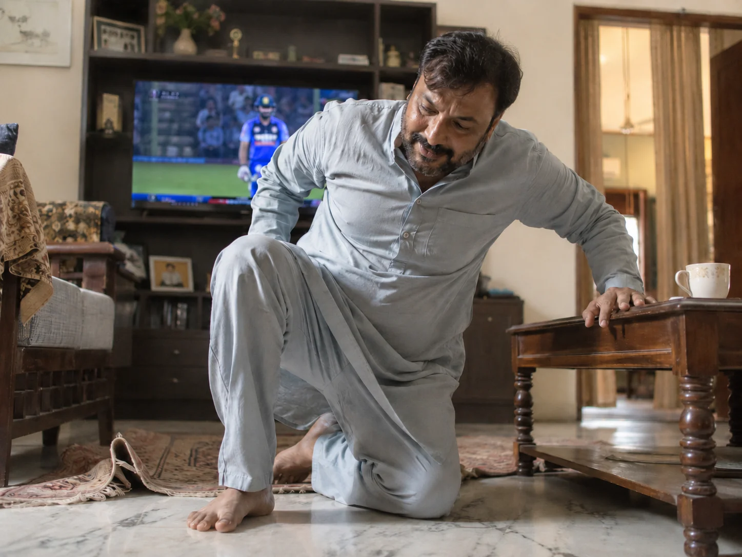 A 43-year-old Indian man using the coffee table to get off the floor — cricket on TV, chai on the table